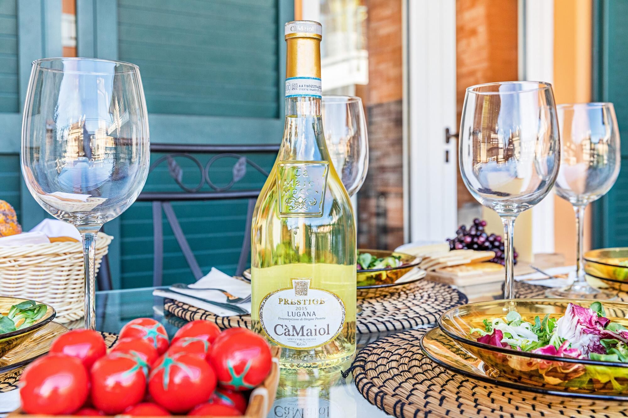 Uma mesa posta com taças de vinho, uma garrafa de vinho Ca Maiol e diversos alimentos, incluindo tomates e uma saladeira.