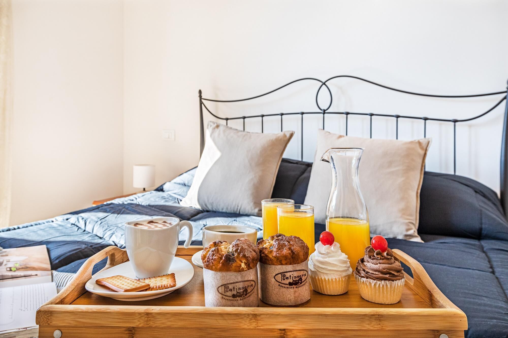 A breakfast tray with pastries, a cup of coffee, juice, and a pitcher on a bed with a metal headboard.