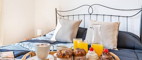 Bandeja de desayuno con pasteles, una taza de café, jugo y una jarrón sobre una cama con cabecera de metal.