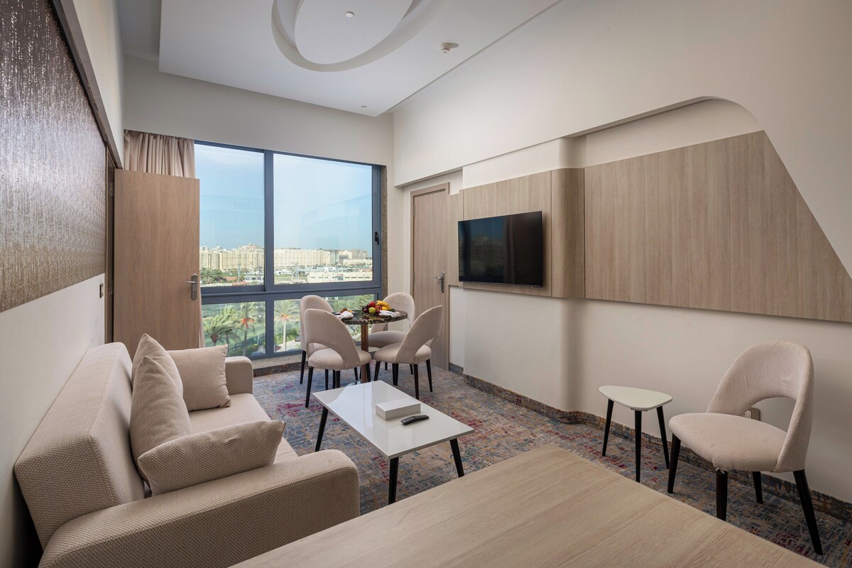 A modern hotel room with a large window, a sofa, a dining table, and a TV mounted on the wall.