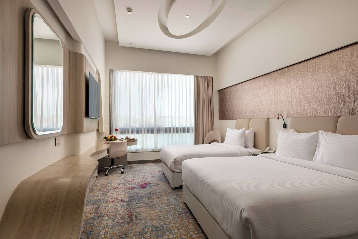 Ein modernes Hotelzimmer mit zwei Betten, einem Schreibtisch, einem Sessel, einem Fernseher und einem großen Fenster mit Vorhängen.