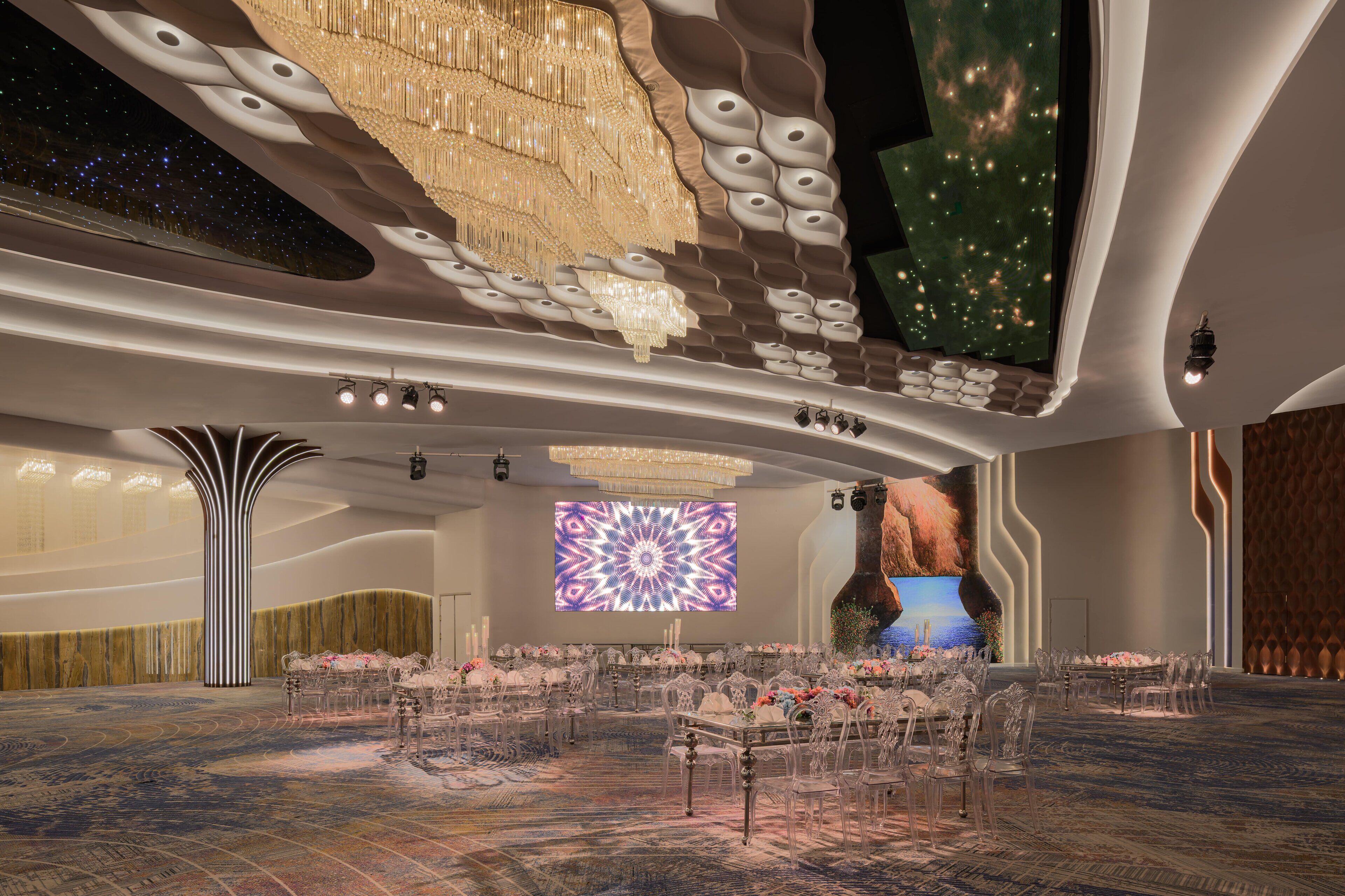 A spacious hall with a high ceiling, modern chandeliers, and a large screen displaying a geometric pattern.