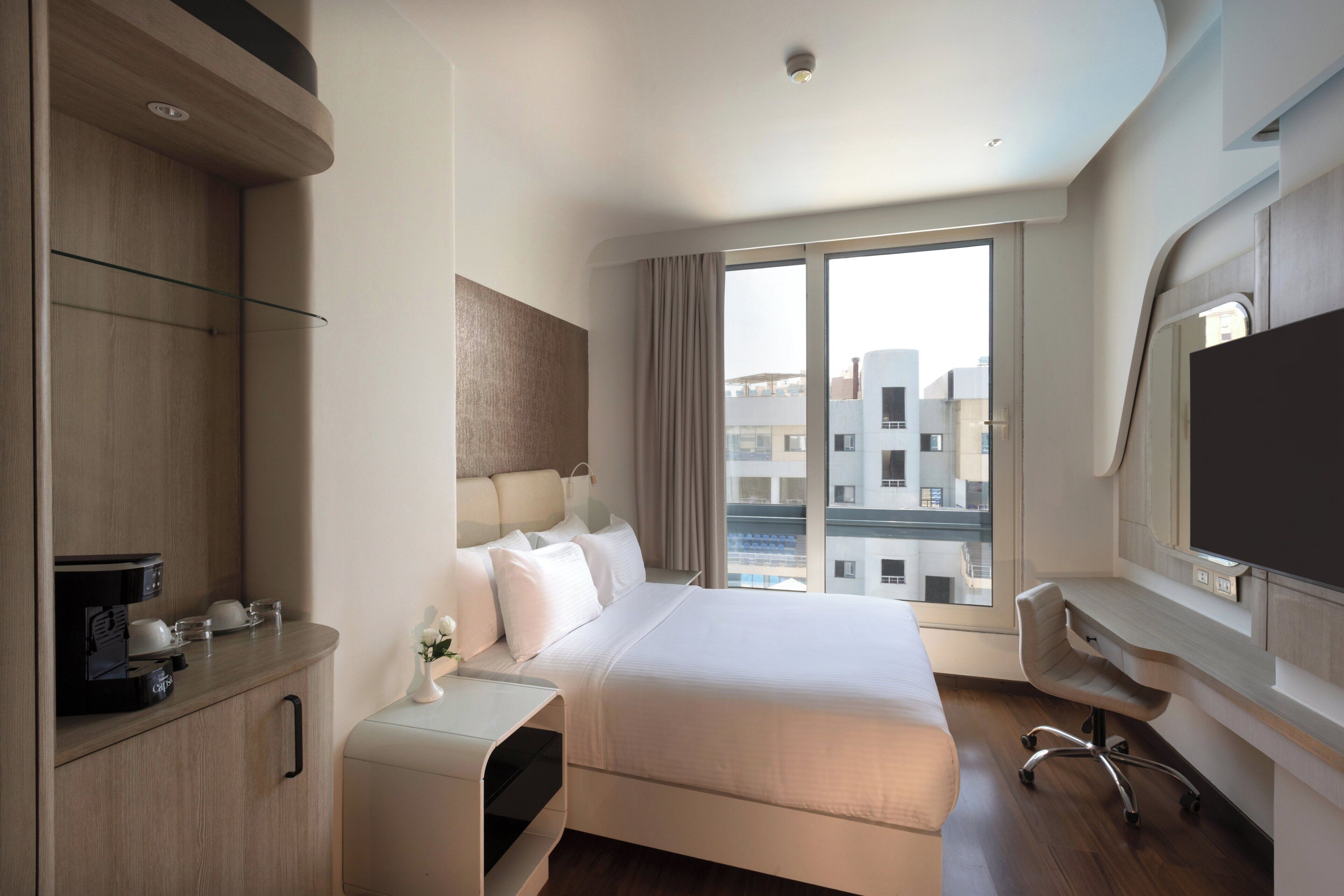 A modern hotel room with a bed, a desk, a chair, a TV, and a window with a city view.