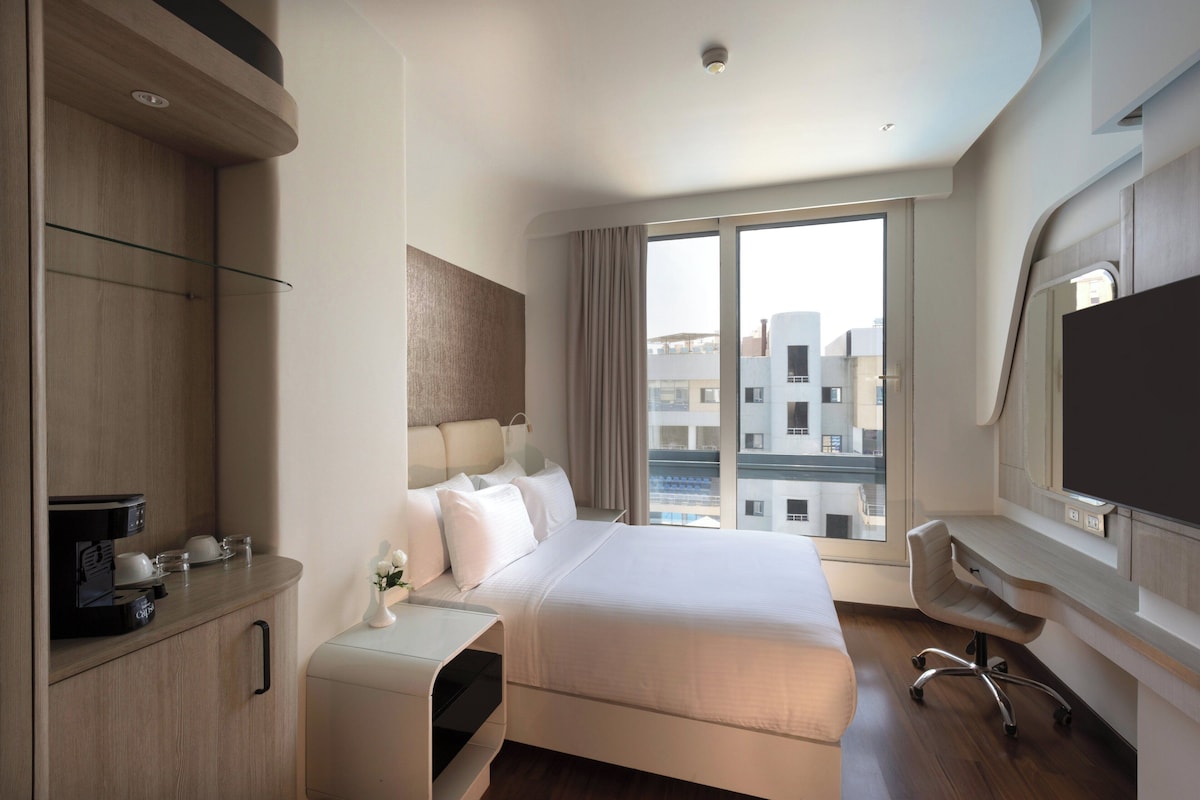 A modern hotel room with a bed, a desk, a chair, a TV, and a window with a city view.