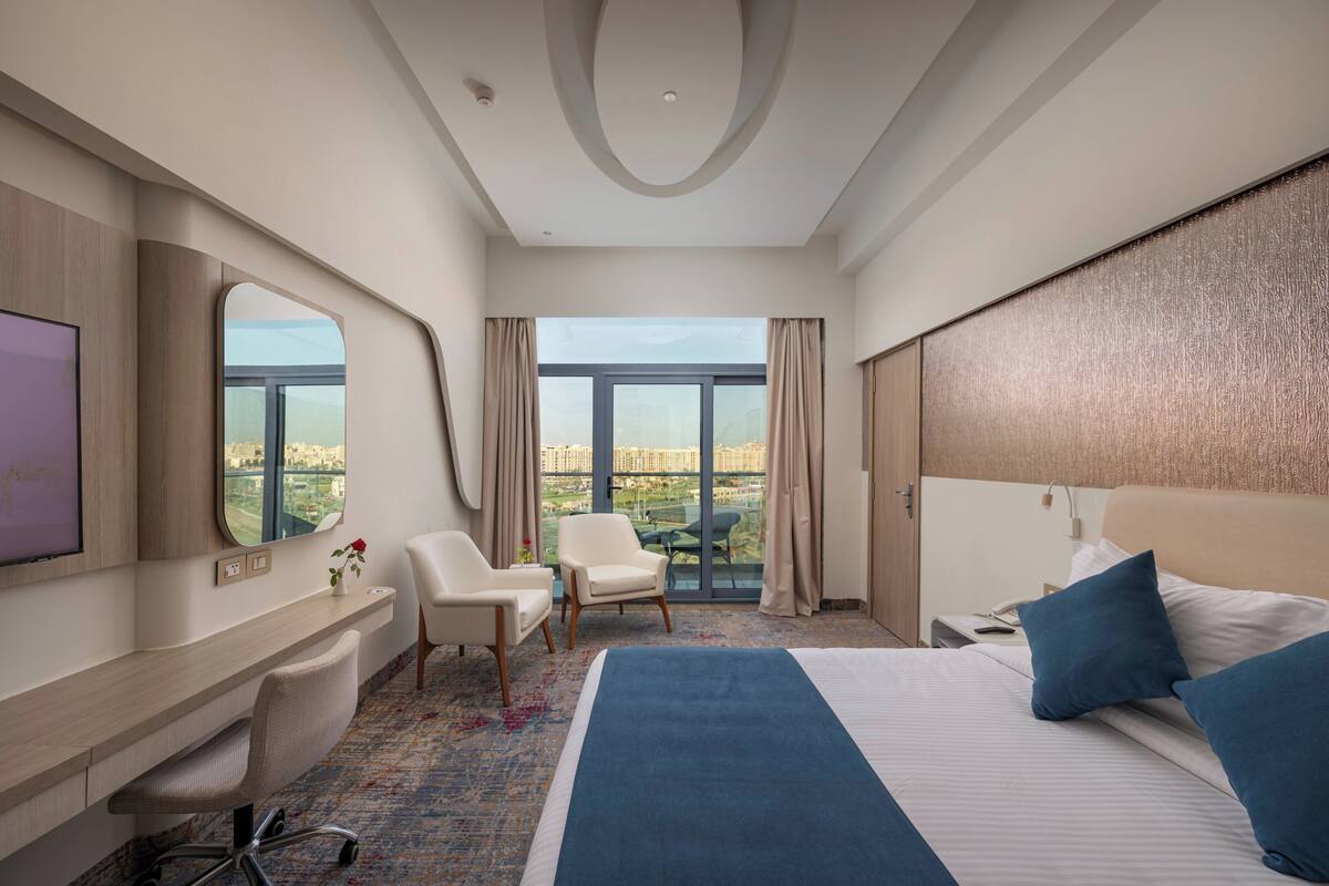Ein Hotelzimmer mit einem großen Bett, einem Schreibtisch, einem Sessel, einem Fernseher und Blick auf die Stadt.