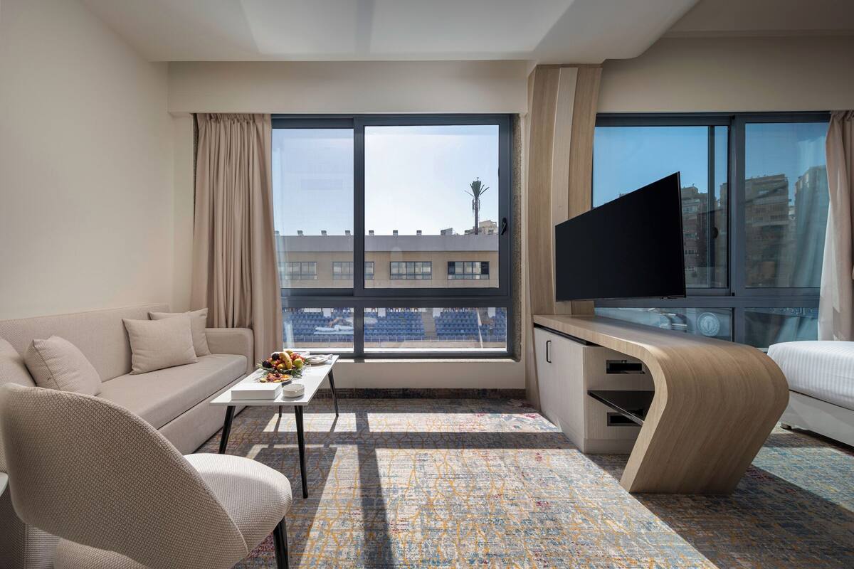 Ein modernes Hotelzimmer mit einer Couch, einem Sessel, einem kleinen Tisch mit Obst, einem Flachbildfernseher und einem großen Fenster mit Blick auf die Stadt.