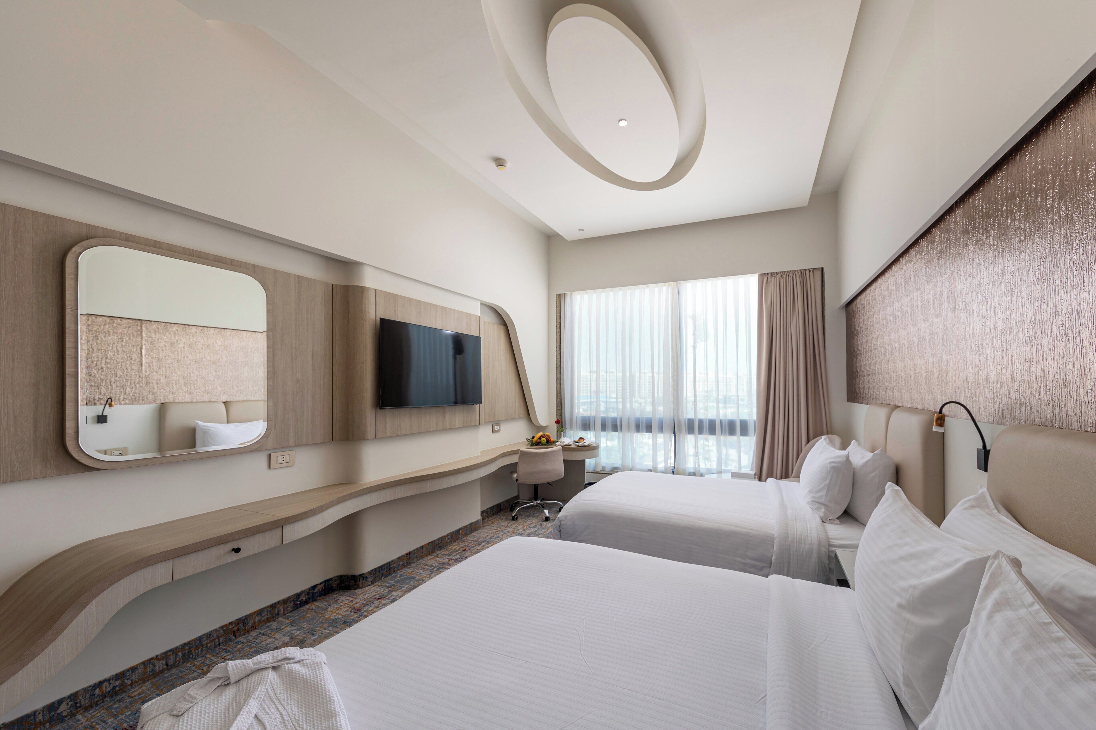 Une chambre d’hôtel avec deux lits, un grand miroir, un téléviseur à écran plat, un bureau avec une chaise et une grande fenêtre avec des rideaux.