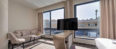 A modern hotel room with a sofa, a small table with fruit, a TV on the wall, and large windows with a city view.
