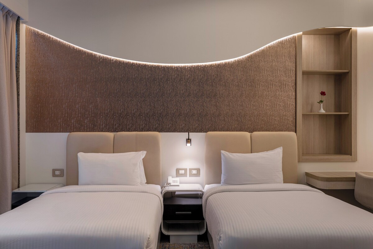 A hotel room with two beds, a curved headboard, a bedside table with a lamp, and a small shelf with a vase.