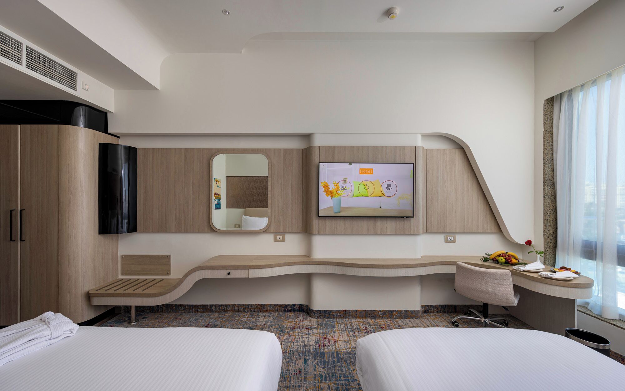 A modern hotel room with a curved desk, two beds, a flat-screen TV, a mirror, and a window with curtains.