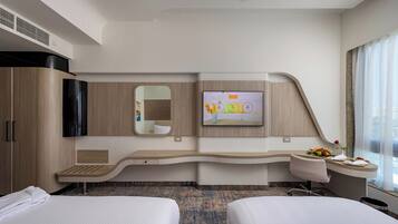 A modern hotel room with a curved desk, two beds, a flat-screen TV, a mirror, and a window with curtains.
