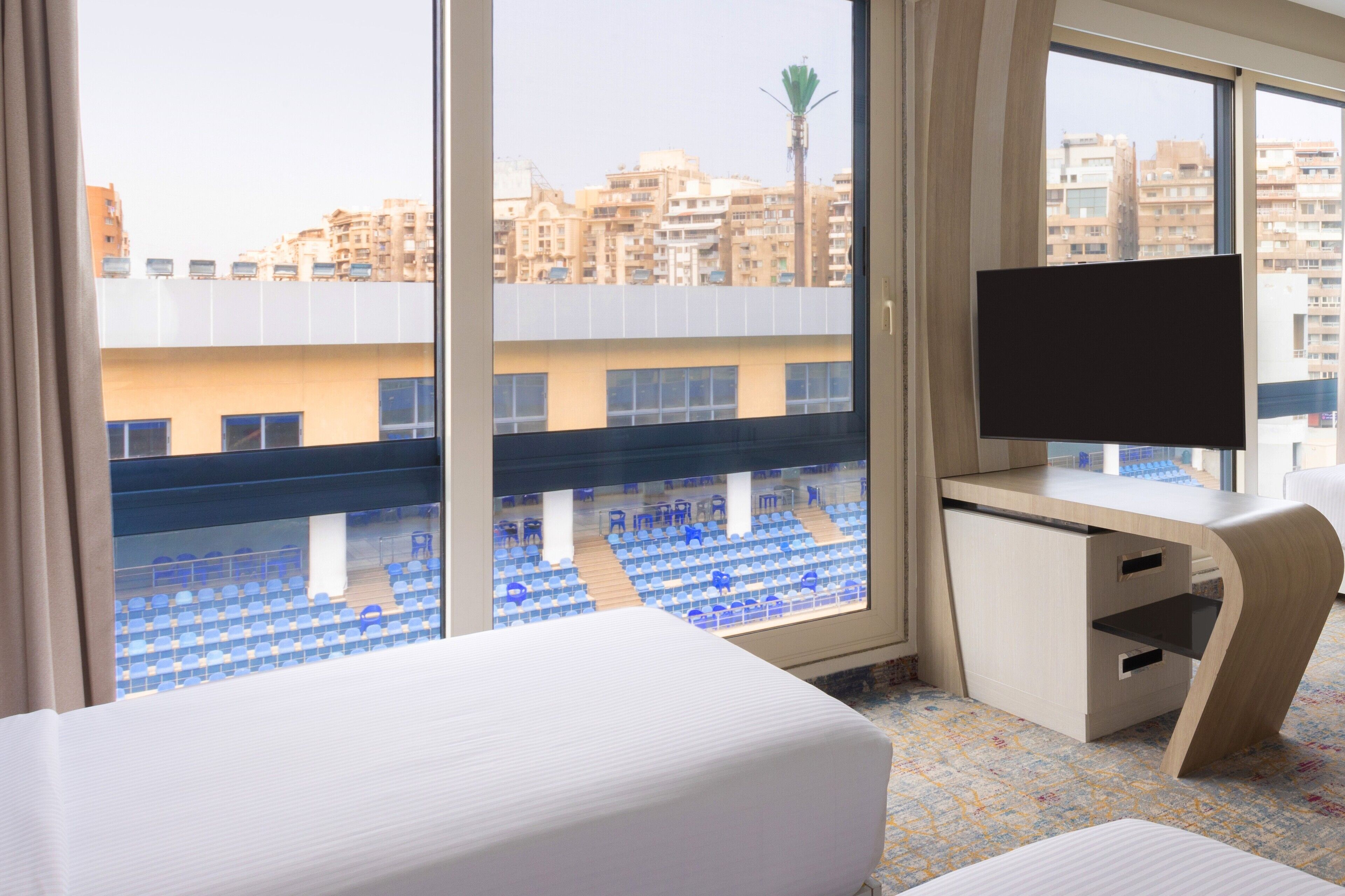 Habitación de hotel con cama, escritorio y televisor, y vista a un edificio con sillas azules en el exterior.