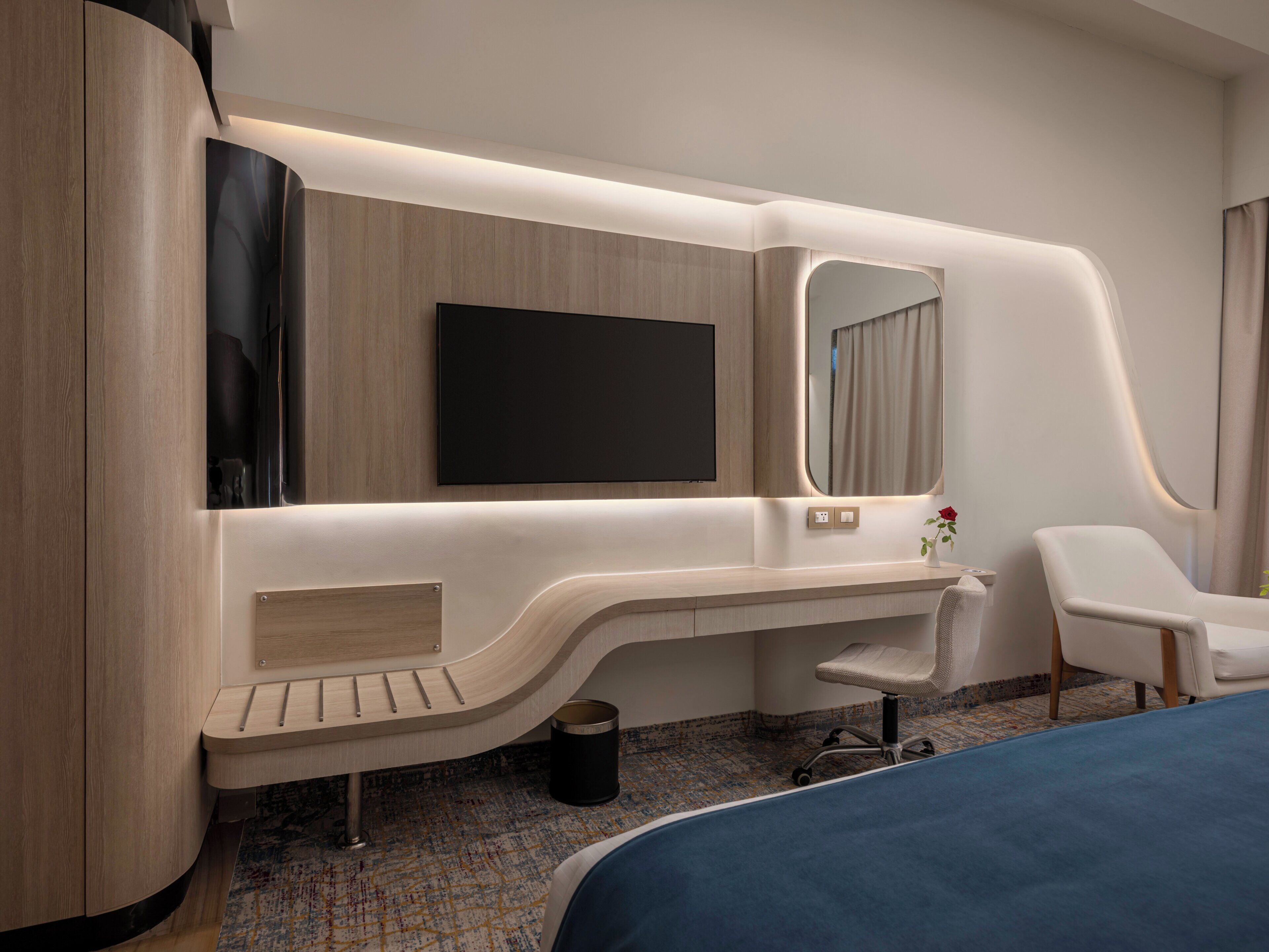 A modern hotel room with a curved desk, a flat-screen TV, a large mirror, a white chair, and a bed with a blue cover.