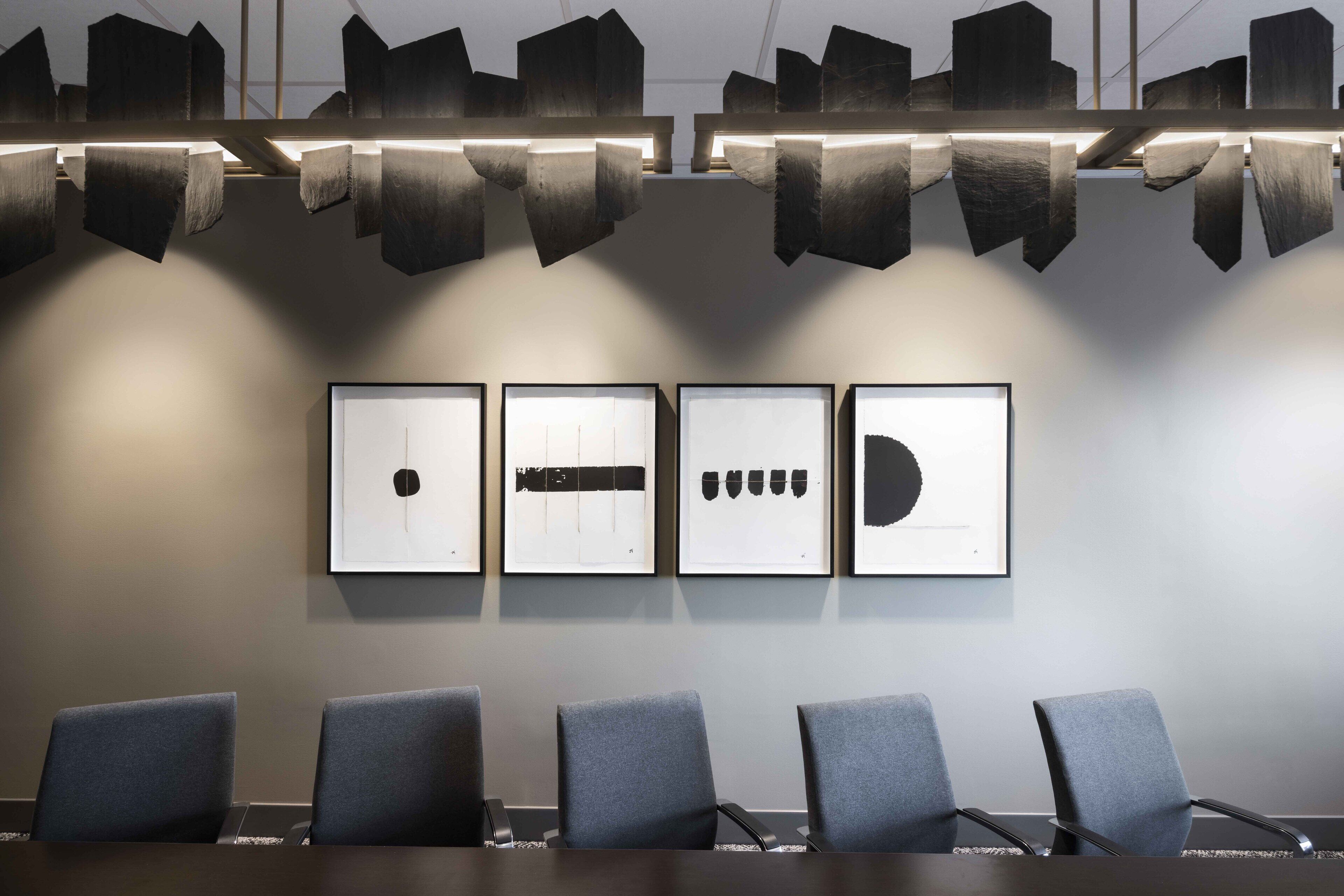 A conference room with a long table and six chairs, four framed artworks on the wall, and a unique ceiling light fixture.
