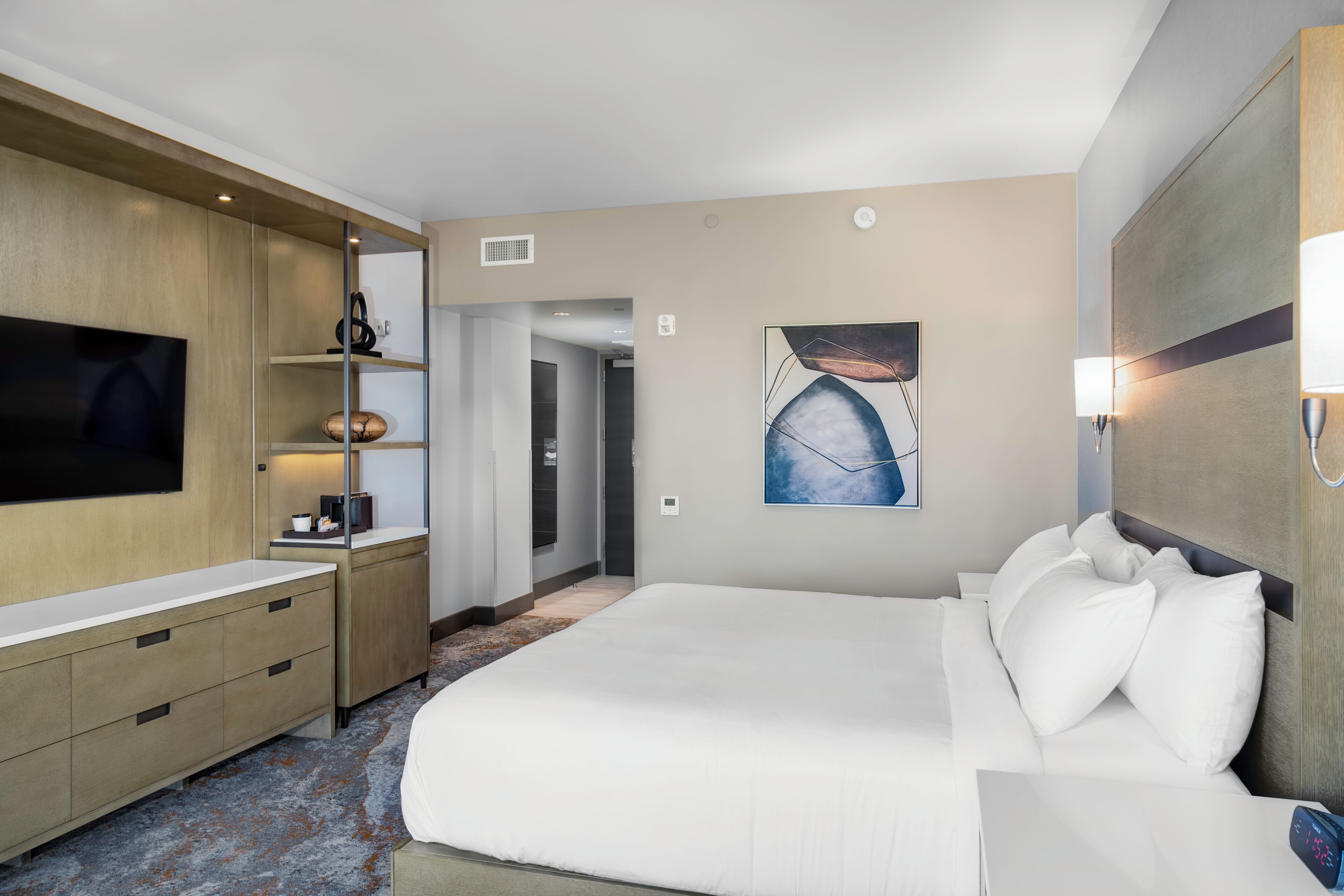 Ett modernt hotellrum med en stor säng, en platt-TV, ett skrivbord med en lampa och en tavla på väggen.