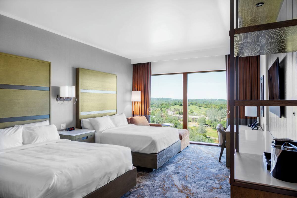 Habitación de hotel con dos camas, un escritorio, una silla, un ventanal con vistas y un mini bar.
