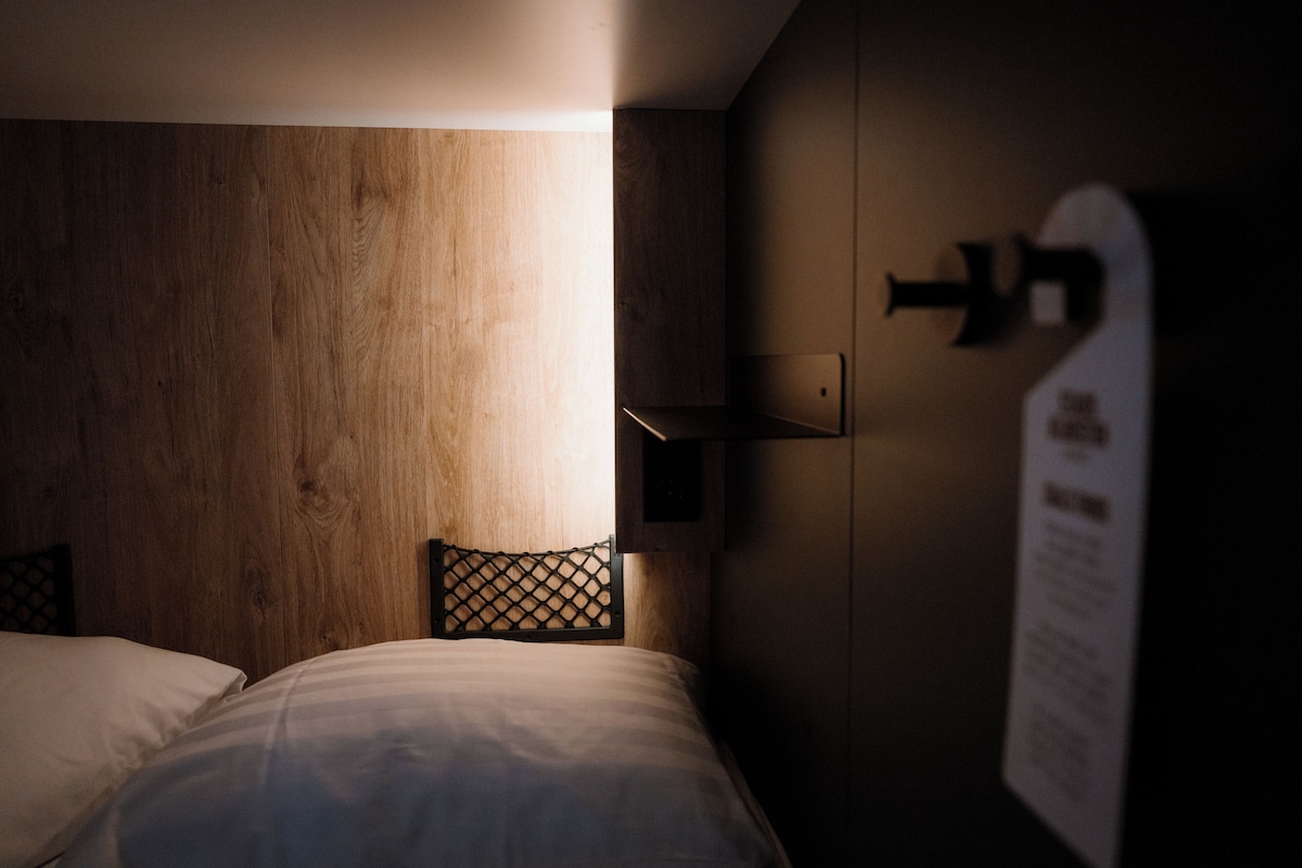 Hotellihuone, jossa on puinen sängynpääty, valkoisilla lakanoiden peitetty sänky, seinään kiinnitetty valaisin ja tekstillä varustettu kyltti.