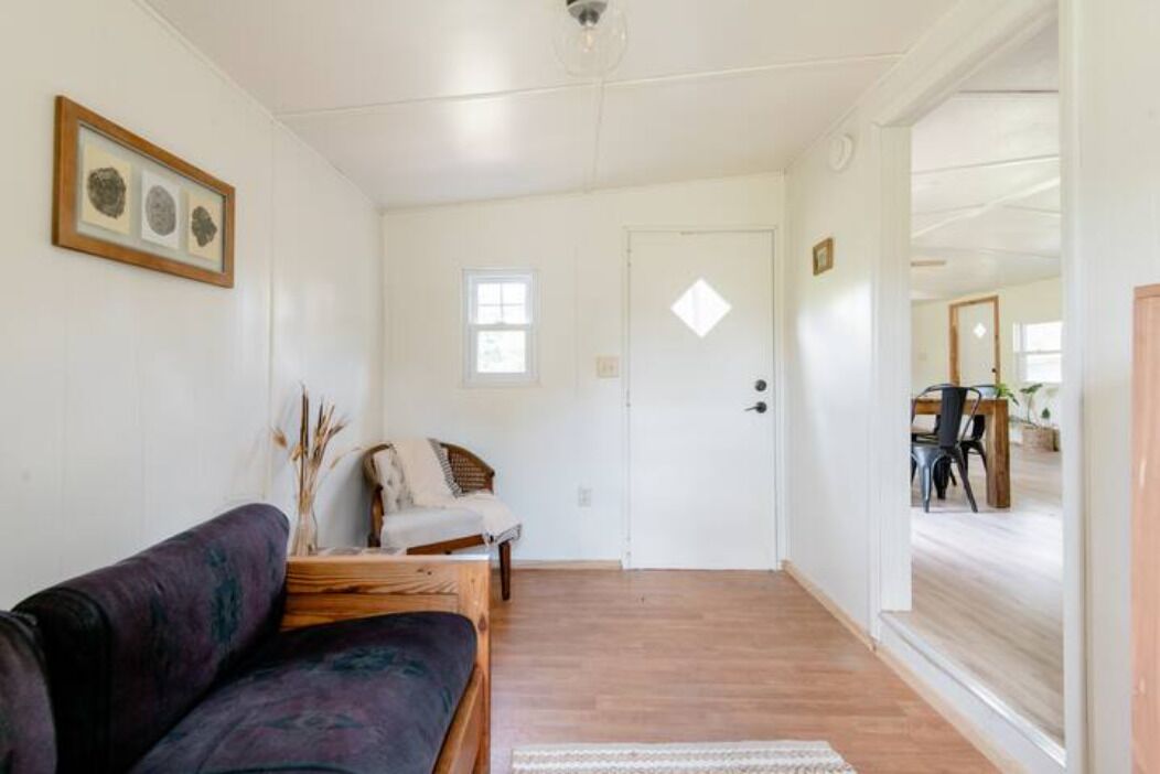 A room with a wooden floor, a sofa, a chair, a small table with a vase, and a door leading to another room.