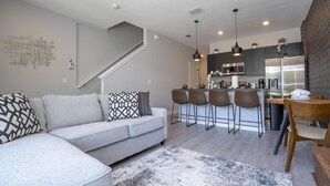 A modern living room with a grey sofa, patterned cushions, a coffee table, and a rug. There is a kitchen area with a bar and seating, and a staircase leading to another level. - 3280 - Beautiful 3br Home near Disney - The Hub at Westside (Kissimmee)