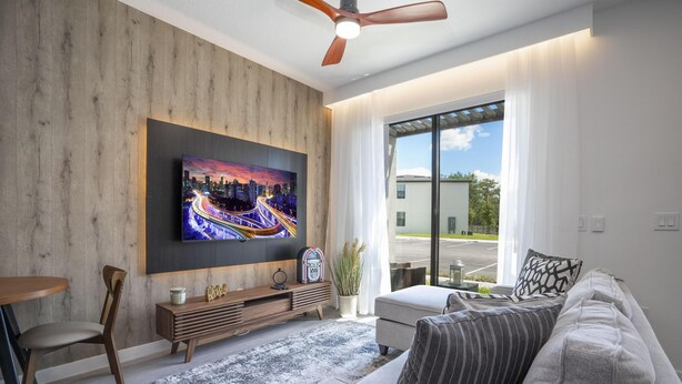 A modern living room with a flat-screen TV mounted on a wooden wall, a grey sofa, a wooden coffee table, and a dining area with a chair and table. - 3280 - Beautiful 3br Home near Disney - The Hub at Westside (Kissimmee)