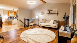 A living room with a sofa, a round rug, a wicker chair, a dining area with chairs, and a kitchen in the background. - Midlake Elegance Retreat (Hudsonville)