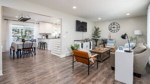 A spacious living area with a dining table, a sofa, and a coffee table. There is a kitchen area with white cabinets and a large window.