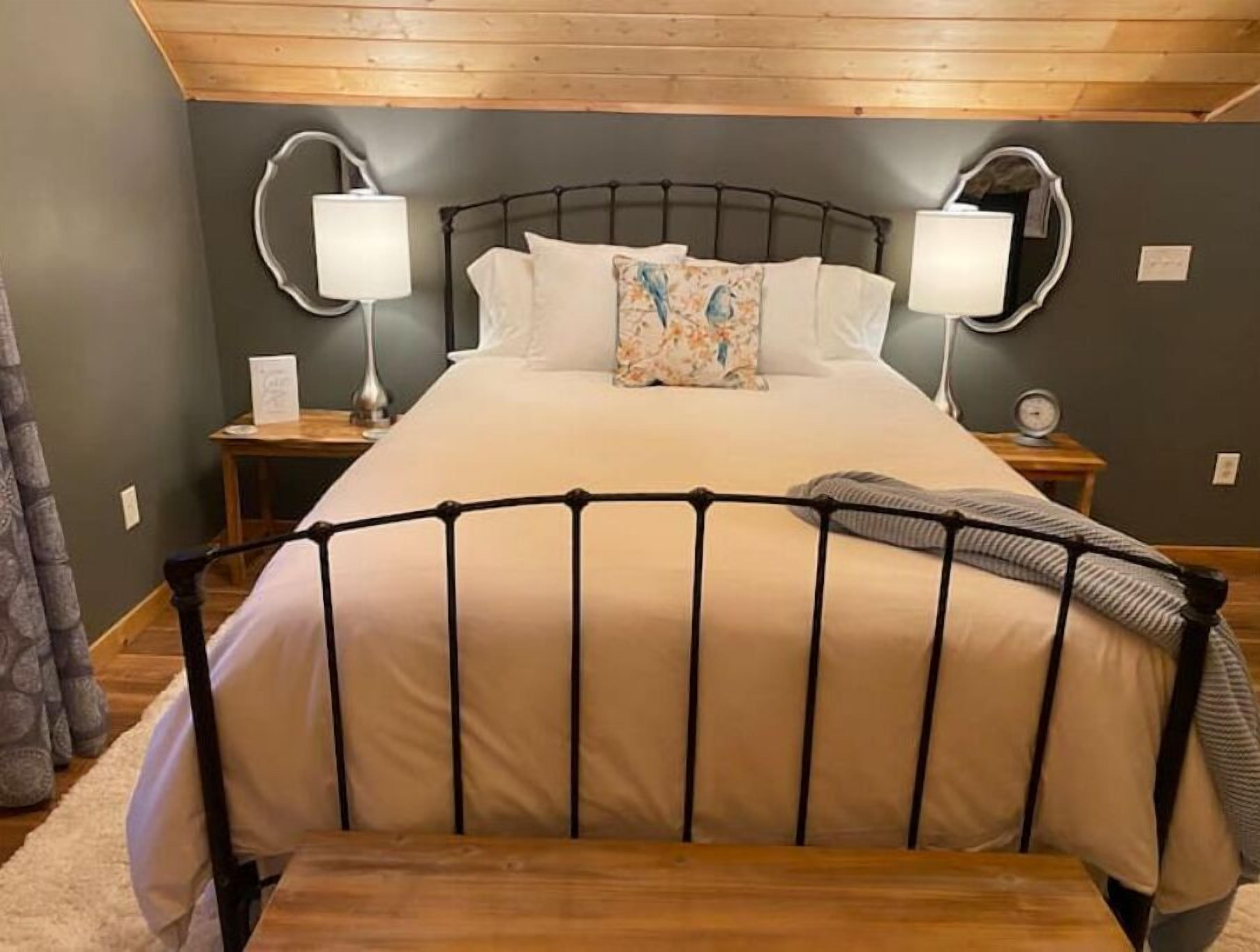 Quarto com uma cama, duas mesas de cabeceira, uma lâmpada em cada mesa e um relógio em uma delas.