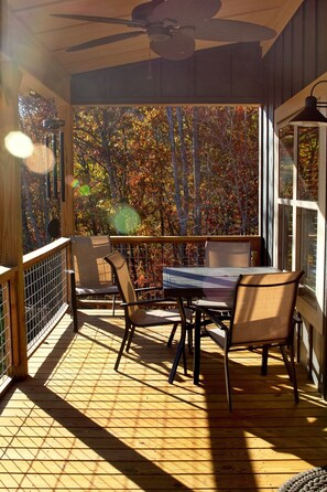 Une terrasse en bois avec une table et des chaises, un ventilateur de plafond et une vue sur les arbres aux couleurs automnales, visible à travers de grandes fenêtres.