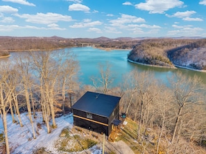 Une cabane dotée d’un toit sombre et d’un petit porche, entourée d’arbres sans feuilles et surplombant un sol enneigé, avec vue sur un lac paisible et des collines lointaines.