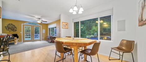 Un comedor amplio con una gran mesa de madera y cuatro sillas, un ventilador de techo y ventanas grandes que ofrecen vistas al exterior.