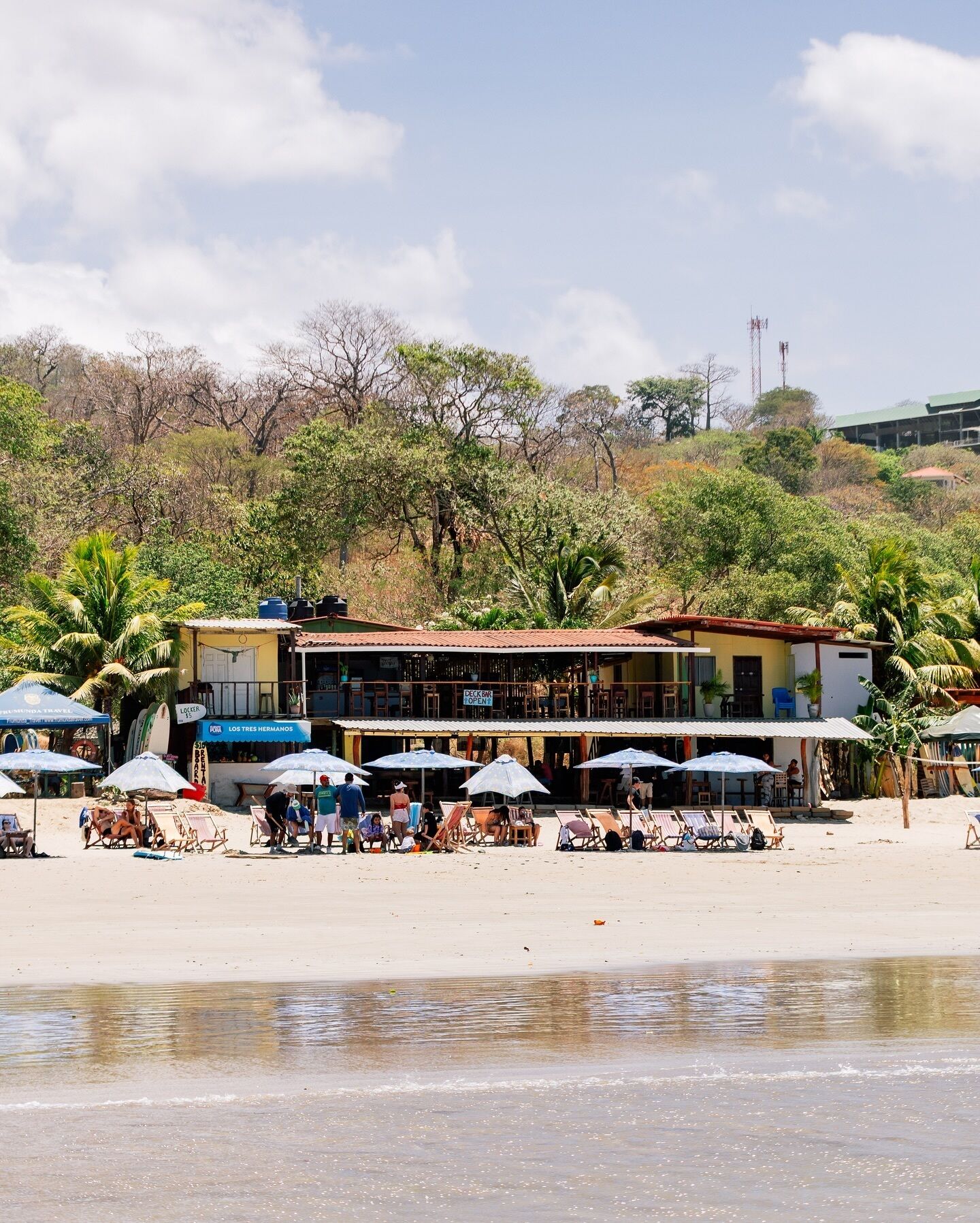 A beachside building with multiple umbrellas and lounge chairs, a sign indicating 'DECK OPEN', and a view of the beach and trees in the background.