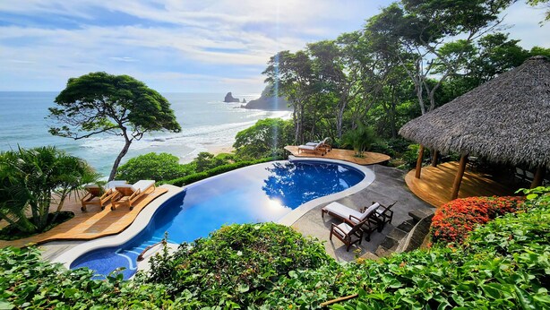 A pool with a view of the ocean, surrounded by lush greenery and a thatched-roof structure. - BEACHFRONT INFINITY POOL – DIRECT PRIVATE BEACH ACCESS – FOREST & TROPIC GARDENS (San Juan del Sur)
