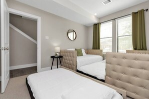 Two beds with tufted headboards, a round mirror, and a side table with a lamp.
