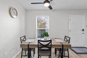 A dining room with a wooden table, four chairs, a ceiling fan, a clock, a window with blinds, and a door.