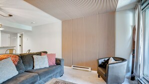 A modern living room with a grey sofa, a coffee table, and a black armchair. There is a wooden slatted wall behind the sofa and a window with curtains on the right. - Walk to Ski Lift + Hot Tub: Copper Mountain Condo (Frisco)