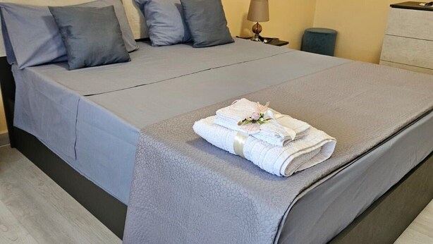 A neatly made bed with a grey bedspread and pillows, a folded towel with a flower, a bedside lamp, and a dresser. - ROYAL HOUSE PALACE (Caserta)