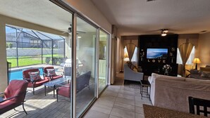 A spacious balcony with a glass door leading to a pool area, a living room with a TV and seating, and a view of a landscaped yard. - Solara Resort private POOL HOME 4 Bed (Kissimmee)