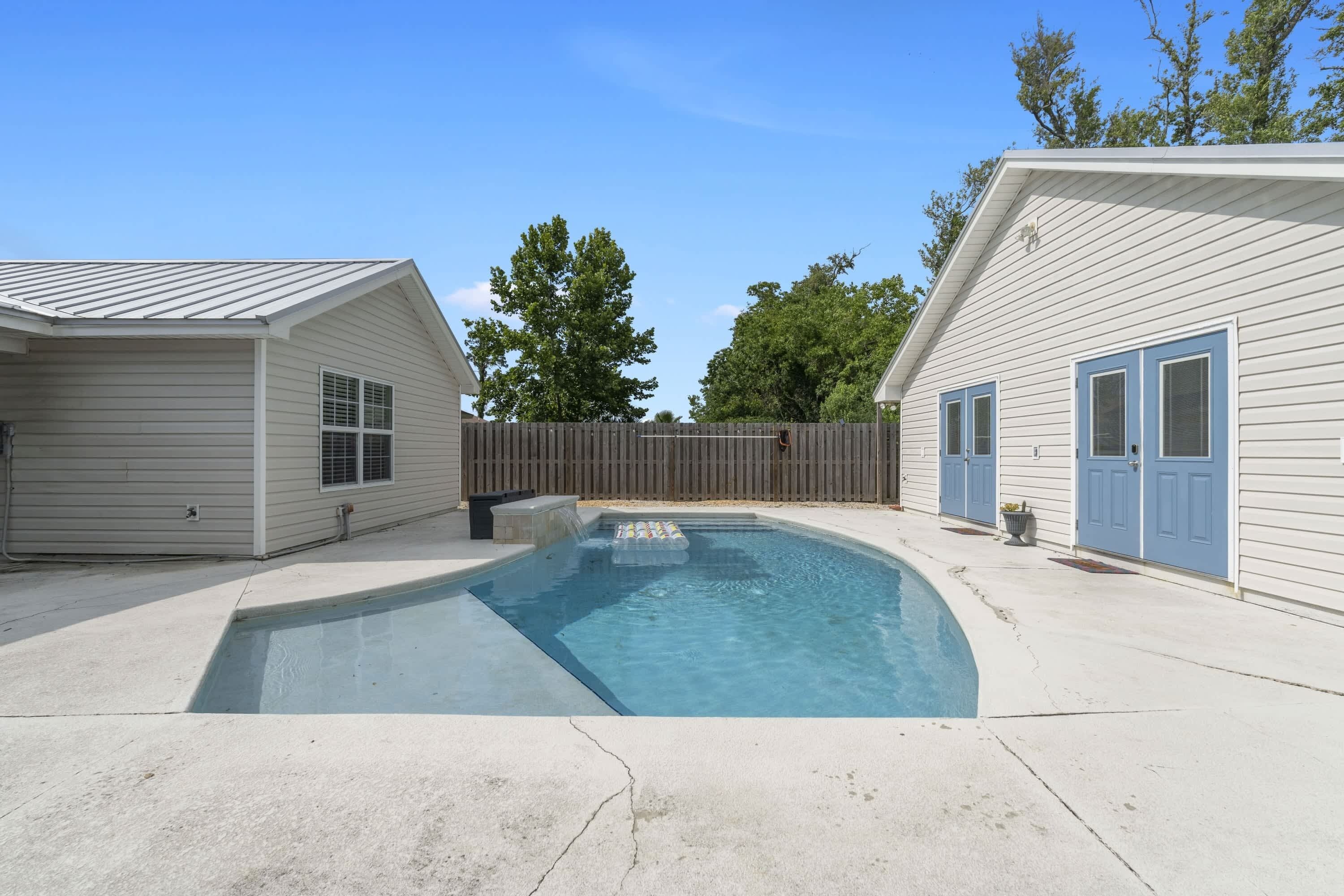 A swimming pool with a wooden deck, surrounded by two buildings with blue doors and a wooden fence.