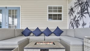 A covered outdoor seating area with a fire feature, a light-colored wall, and a window.