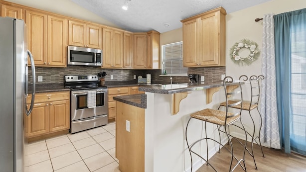 A modern kitchen with wooden cabinets, stainless steel appliances, a granite countertop, and a breakfast bar with metal chairs.