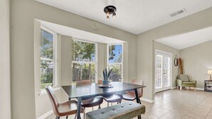 Un espace repas comprenant une table, des chaises et un banc. Il y a des fenêtres avec des stores, un luminaire au plafond et une porte donnant sur l’extérieur.