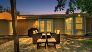 Une terrasse couverte avec une table et des chaises, un barbecue et une maison avec un portail de garage.