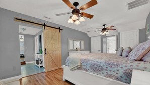 A bedroom with a large bed, wooden flooring, a ceiling fan, and a sliding barn door. - Spacious Family Retreat with Game Room 25 Mins to PCB (Lynn Haven)