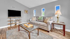 A living room with a gray sofa, a coffee table, a TV on a stand, and a lamp on a side table. - Adorable 1 BDR with Full Kitchen Deck Washer Dryer (Lynn Haven)