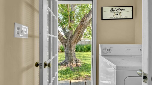 Una sala de lavandería con lavadora y secadora, una puerta que da a un patio con un árbol grande, y un cartel en la pared que dice "Calcetines perdidos buscando su media perfecta."