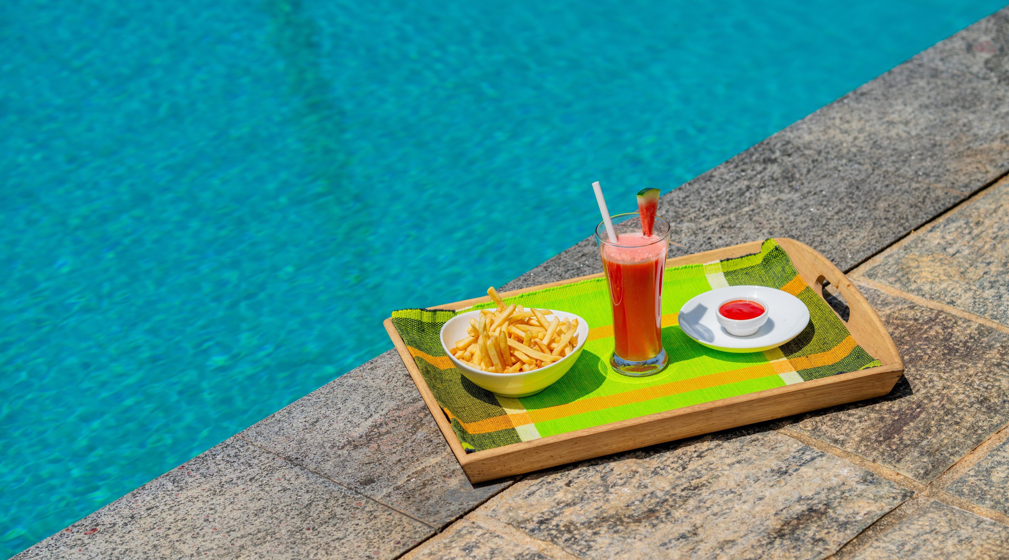 A tray with a bowl of fries, a glass of red drink, and a small dish of ketchup by a poolside.