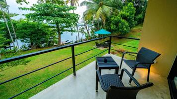 A balcony with a table and chairs, overlooking a grassy area with trees and a body of water.