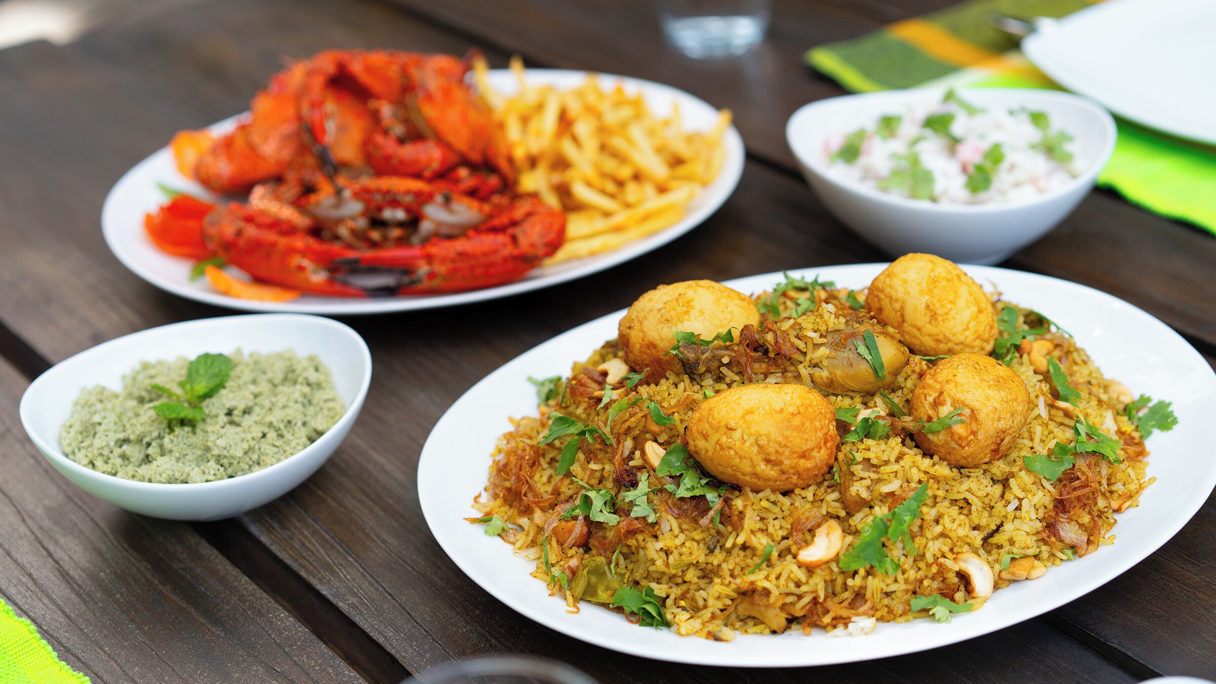 A table with various dishes including a plate of biryani with eggs, a plate of fried seafood, and a bowl of green chutney.