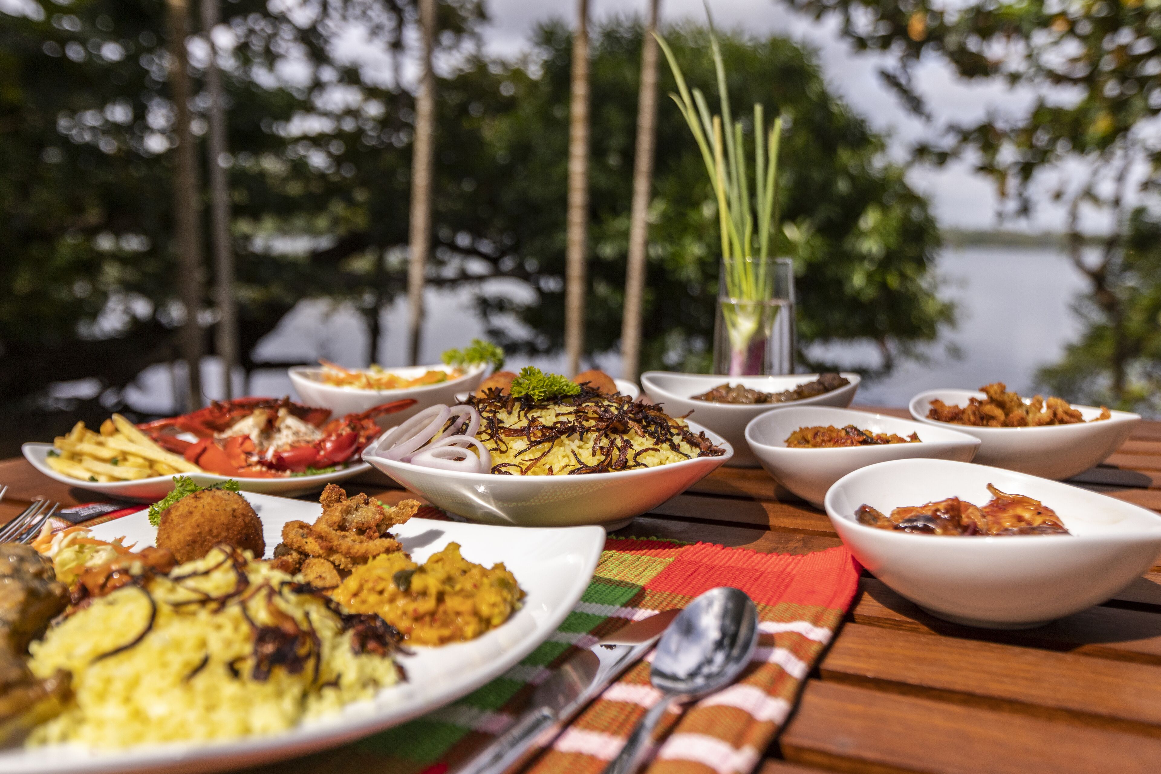 A table set with various dishes, including seafood and rice, with a view of trees and water in the background.
