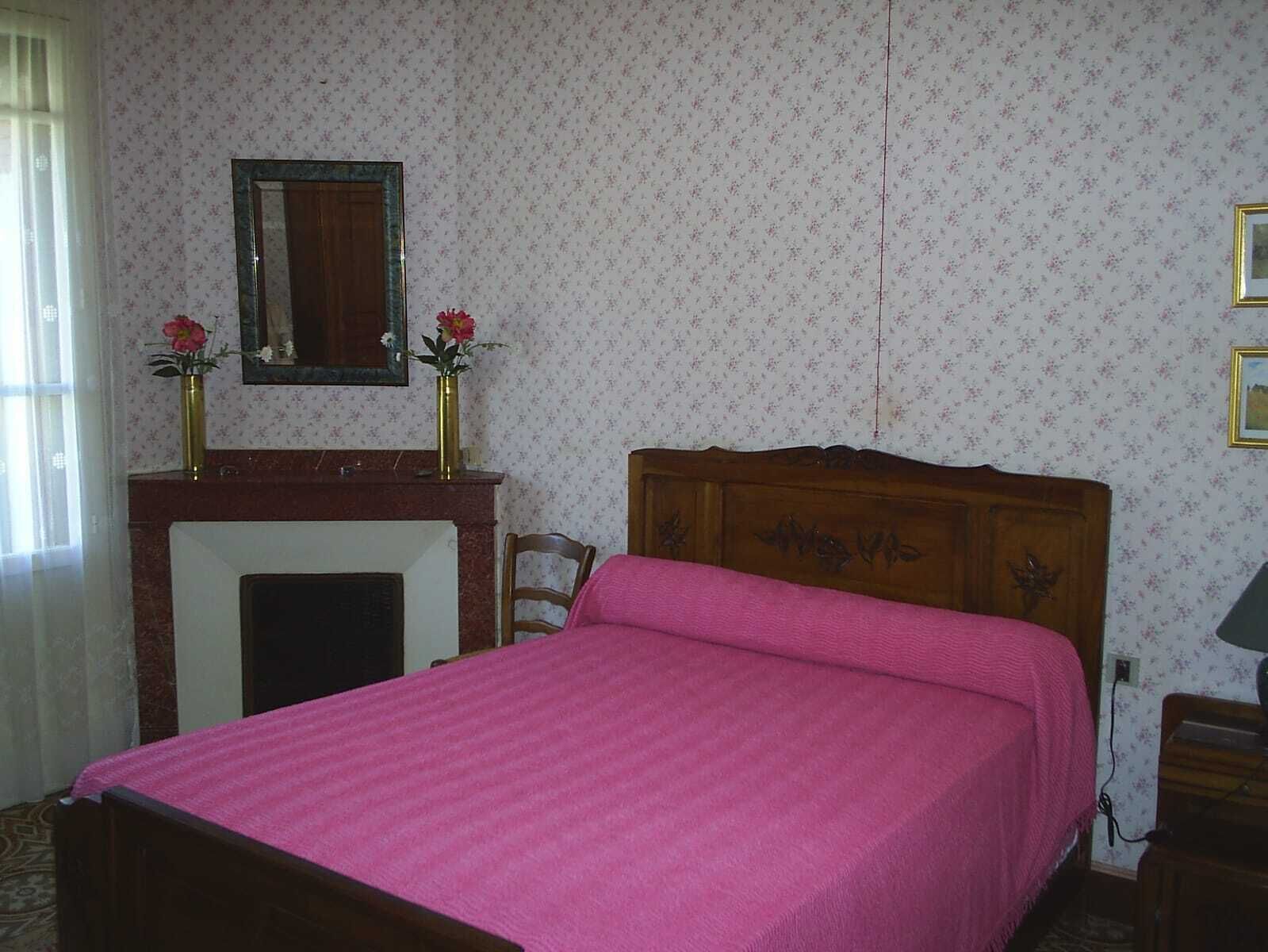 A bedroom with a wooden bed, a fireplace, floral wallpaper, and a mirror above the mantel.
